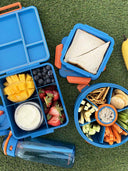 Blue lunch box with compartments filled with snacks and a sandwich on a grass background