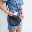 Fleur Saddle Leather Crossbody Handbag Black — Shoulder carry by women in denim dress inserting phone into handbag | Spixal