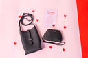  two leather black bags and a card saying "because of her" card behind and a pink background and hearts