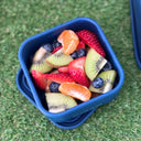 A blue square silicone container filled with sliced fruit, including strawberries, kiwi, blueberries, and orange segments, placed on green grass.