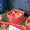 Orange Spixal silicone lunch containers arranged on grass with fruit, a sandwich, and snacks, with a dark gray bag and blue backpack in the background.