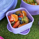 A purple square lunch container filled with sliced strawberries, blueberries, kiwi, and orange segments, placed on grass next to a purple backpack. The container has the brand name 'spixal' printed on the side.